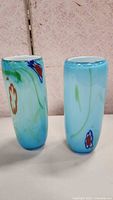 Two light blue cylindrical art glass vases with floral designs on a light pink textured background.
