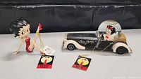 Photo showing the Betty Boop figurine sitting in a posed leg-raised position next to the black and cream vintage-style car with snow globe on top. Both items have attached tags.