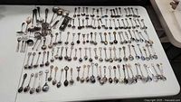 Full view of the large collection of assorted collector spoons and serving utensils arranged on a white table.