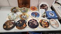 Collection of 12 decorative plates with varying artistic designs including children and literary themes laid out on table