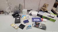 Photo showing various electronics and travel accessories, including a projector, camera charger, pouches, and other small gadgets.