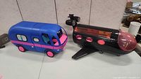 Shows side view of blue and pink camper van and black and pink airplane with transparent cockpit and wings on white table.