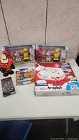 Photo showing all items in the lot arranged on a table with a white background, including Mrs. Incredible figure, two Peppa Pig boxed sets, an Artsplash paint set box, and two small boxed toys