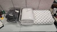 Photo of three bedding items on a table: black quilted coverlet sealed in plastic, off-white queen sheet set in packaging with handwritten note, and white fleece blanket with black polka dots.