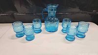 Vintage blue glass lemonade jug in the center, six matching glasses arranged around it, all on a white surface with a dark background.