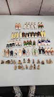 Top-down view of approximately 57 vintage ceramic figurines displayed on a white surface. There are multiple rows of figurines featuring men, women, and children, some holding teapots or cups, colorfully painted with formal and historical costume aesthetics.
