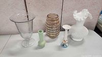 Lot of five vintage vases arranged side by side showing various colors and styles including clear glass, amber swirl, green hobnail, hand painted porcelain, and white hobnail milk glass.