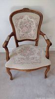 Front view of brocade upholstered wood chair showing detailed fabric design of floral and pastoral scene on backrest and seat, carved wood frame top, arm, and legs.