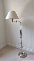 Full view of the adjustable metal floor lamp with white shade against a plain wall and carpeted floor.