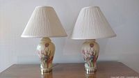 Two identical ceramic floral design lamps with white pleated shades side by side on a wooden dresser.
