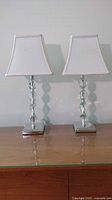 Front view of two matching lamps with crystal-style clear segmented bases and white square lampshades, placed on a wooden surface.