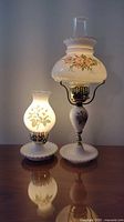 Photo of both milk glass lamps together, one smaller, one larger, both lit showing rose floral pattern.