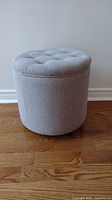 Front view of round grey fabric storage ottoman with tufted cushioned top and smooth sides.
