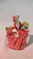 Front view of Lydia porcelain figurine in red dress and hat, seated with book and floral background.