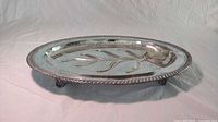 Side angled view of the oval silver plated meat platter showing rope edge, tree shaped juice well, and three small feet.