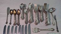 Top view shot showing the assortment of silver plated forks, spoons, and serving pieces arranged horizontally with visible ornate and intricate embossed Floral/scroll handles, some items show tarnish.