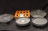 Five plant caddies grouped: black plastic, wooden slat square, clear rimmed, two gray matching