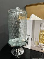 Full view of glass beverage dispenser on pedestal beside box