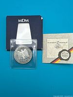 Coin in plastic sleeve alongside MDM box and certificate