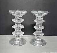 Front view of two clear textured glass candle holders side by side on a light surface with dark background.