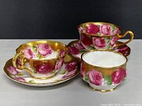 Frontal view of two teacups on saucers and a sugar bowl, showing ornate rose design with gold trim.