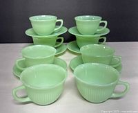Eight pale green Fire King Jadeite cups stacked with six matching saucers beneath, showing overall set composition and condition.