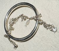 Pair of silver bracelets: a smooth solid bangle and a charm bracelet with fish and leaf-shaped charms and round beads, on a light fabric background.
