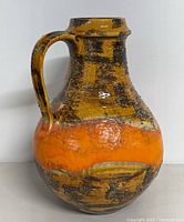Front angle showing textured fat lava glaze jug with handle, predominantly orange with brown and mustard textured areas.