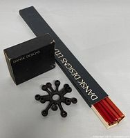Photo showing one box of DANSK DESIGNS branded boxed red taper candles and a starburst design metal candle holder with 10 arms.