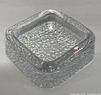 Top angled view of square clear textured glass ashtray with rounded edges and red and white label.