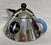 Side angled view of the Alessi modernist teapot with visible Alessi logo sticker, light blue handle with red accents, and black lid knob.