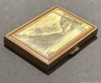 Top view showing the brass compact with scenic engraving on lid depicting mountains, lake, and forest labeled 'Lake Louie Alberta'.