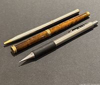 Set of three pens on black background: fountain pen with brown marbled barrel and gold trim, mechanical pencil with wooden barrel and metal accents, and silver Zebra M-402 ballpoint pen.