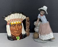 Both ceramic figurines displayed side by side showing front view of detailed Native American chief character jug and seated woman figurine