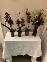 Full view of the four artificial floral arrangements on white draped table showing different orchid plants and mixed bouquet.