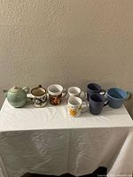 Full set of 8 assorted mugs and cups lined up on white fabric with beige wall background.