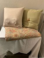 Three items displayed: beige massage pillow, tan pillow with button, and decorative paisley patterned blanket folded beneath.