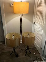 Full view of the floor lamp switched on and two matching table lamps with beige shades and bronze metal bases on a floor.