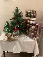 Photo of Christmas decoration lot featuring a faux green pine tree, ceramic tree, box packs of colorful glass ball ornaments, and gold table settings.