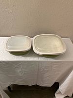 Two square Corelle Vitrelle dishes with lids, olive green exterior with white rims, placed on white cloth against wall.