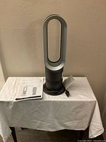 Dyson AM09 bladeless fan and heater unit shown fully assembled with power cord coiled on base and operating manual on white cloth-covered table.