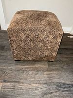 Front view of the square ottoman showing patterned gold and brown upholstery on wood flooring.