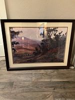 Framed landscape print with wooden frame and visible cream-colored mat and brown accent. Shows trees and hills in the landscape.