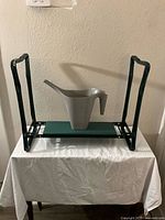 Green metal plant stand with green shelf, grey plastic watering can placed on it, shown indoors on a white cloth-covered surface.