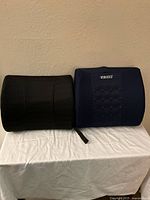Two lumbar support cushions side by side on white cloth, black fabric and blue mesh cushions visible with elastic strap on black cushion