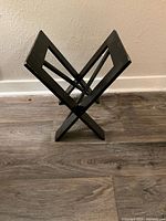 Front view of black wooden geometric magazine/newspaper stand showing the 'X' frame style and angled slots for storage.