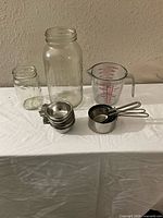 Wide photo showing two glass mason jars without lids, a glass measuring cup, and sets of stainless steel scoops and measuring cups on a white cloth surface against a beige wall.