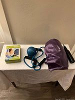 Photo showing all exercise equipment items on white covered table including purple resistance band, blue ball with tubing, black tubular item, and boxed Gaiam Pilates Bodyband.