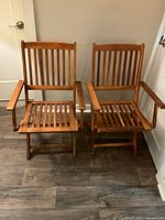 Front view of two medium brown wooden folding chairs with slatted backs and seats, armrests, positioned side by side on dark wood flooring.