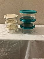 Full set of Pyrex glass storage containers stacked, 3 smaller with white lids on left, 3 larger with teal lids on right.